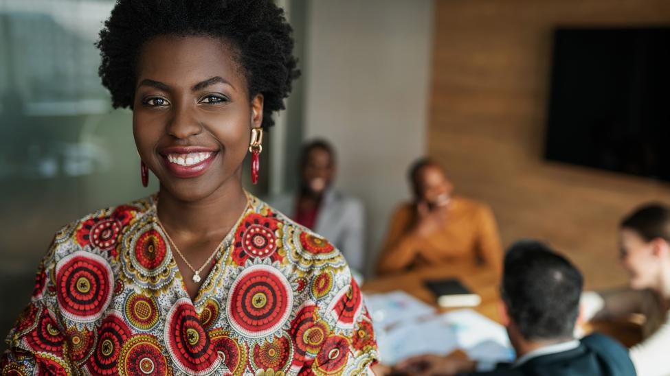 Close up of young smiling professional african woman, team members hold a meeting in background